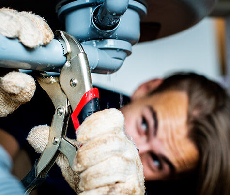 A close-up of a person using a wrench to fix a pipe under a sink. The individual, wearing gloves, is intensely focused on the task as water visibly leaks from the pipe—a scene that highlights the expertise of emergency plumbers in critical situations.