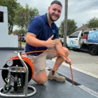 A smiling worker in a blue shirt and beige shorts kneels on the ground and gives a thumbs up. He holds an orange hose that is connected to a piece of equipment on the left. A company van, showcasing Plumbers Sydney, is parked in the background on a suburban street.