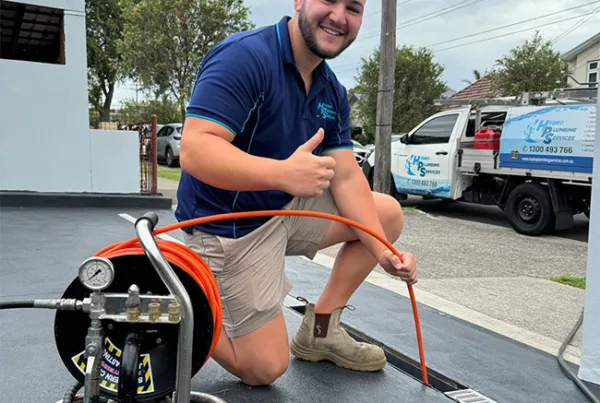 A smiling worker in a blue shirt and beige shorts kneels on the ground and gives a thumbs up. He holds an orange hose that is connected to a piece of equipment on the left. A company van, showcasing Plumbers Sydney, is parked in the background on a suburban street.